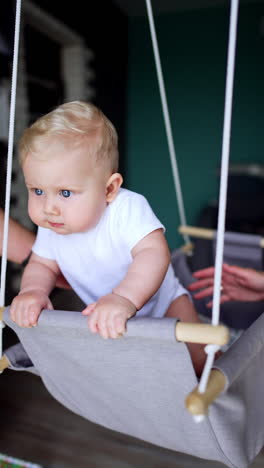 sweet caucasian baby boy in white t-shirt sits in a swing at home. active child tries to stand up and parents support him. vertical video.