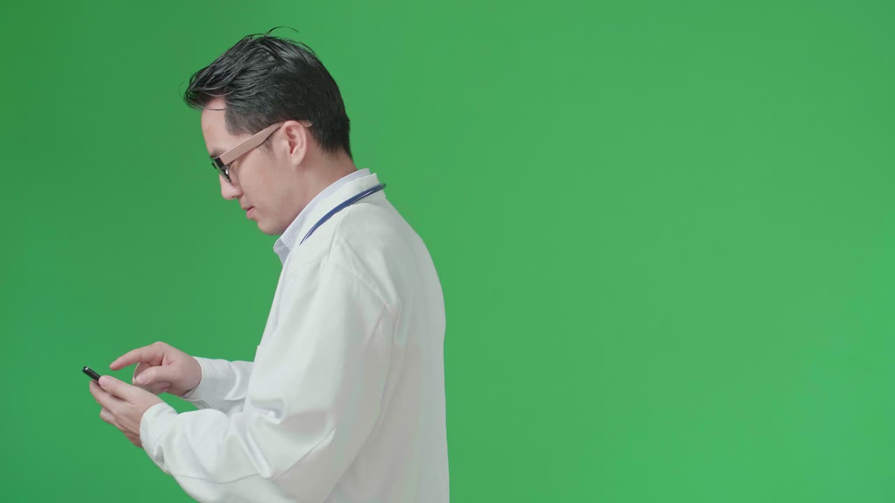 The Side View Of Asian Doctor With Stethoscope Wearing Glasses And White Coat Using Mobile Phone While Walking On Green Screen Chroma Key.