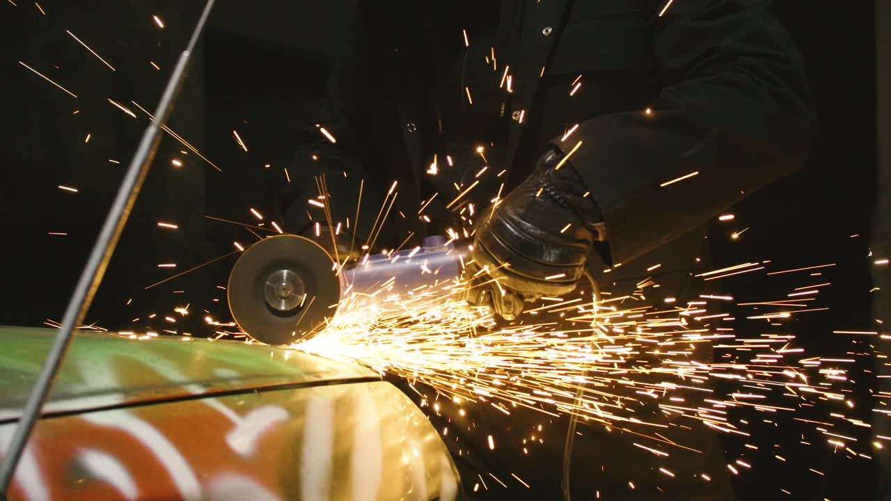Destroying junk graffiti covered car in dark workshop with grinder, sparks flying
