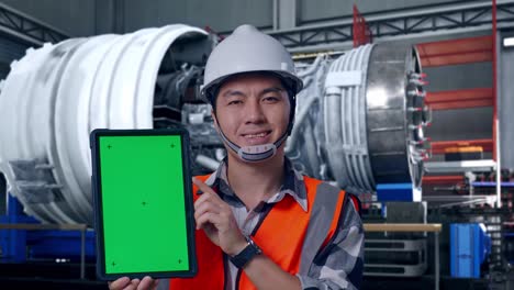 close up of asian male engineer with safety helmet smiling and showing green screen tablet to the camera while standing with airplane engine maintenance conducted, analytics checking the turbine