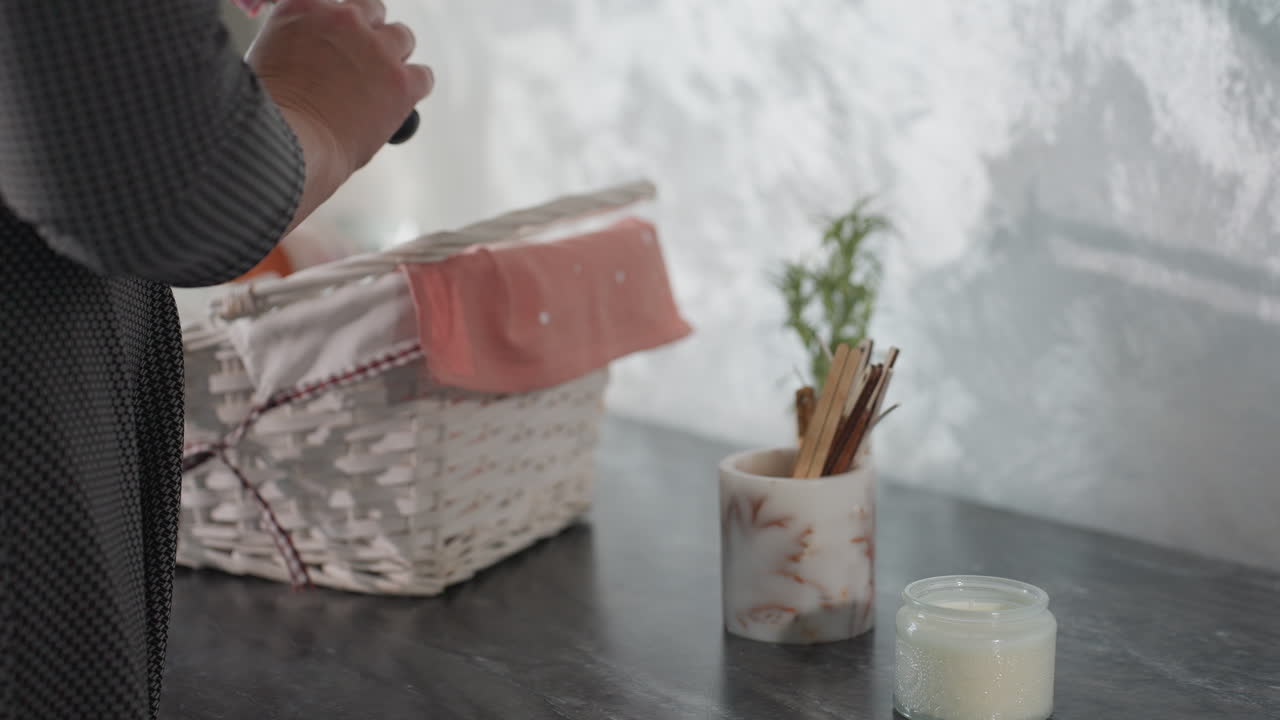 Close up of woman in checkered outfit returning from shopping placing white woven basket with pink cloth liner on black marble table and picking item from inside near candle and potted plant