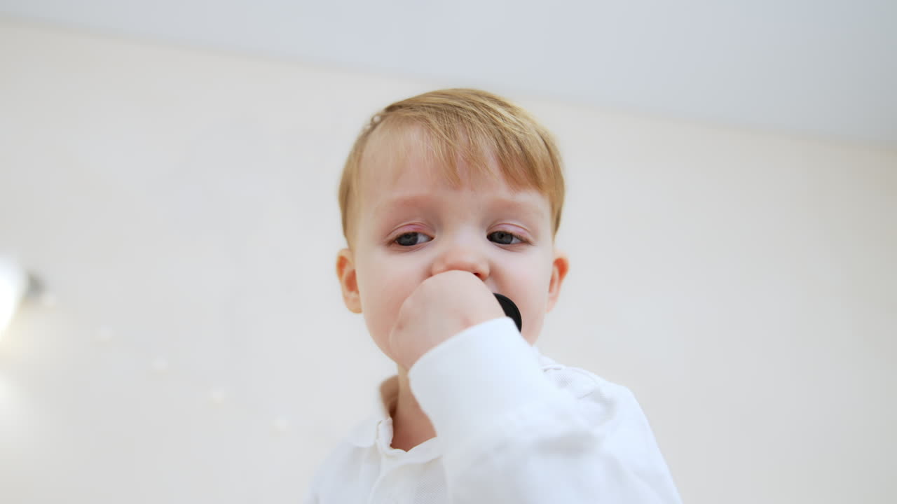 Little Caucasian toddler in white shirt stands looking aside. Kid turns to camera, picks a pacifier and puts it his mouth. Low angle view.