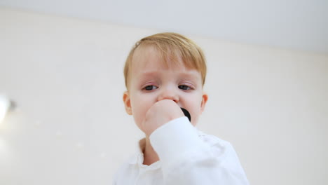 little caucasian toddler in white shirt stands looking aside. kid turns to camera, picks a pacifier and puts it his mouth. low angle view.