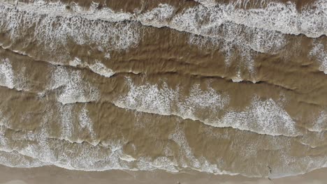 Top-down-stationary-view-of-the-waves-crashing-with-people-in-the-water-in-the-bottom-right-corner