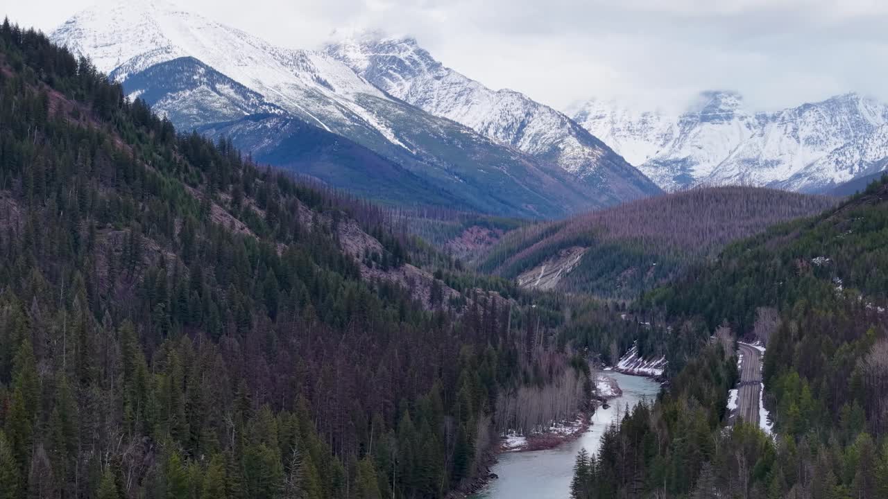 Vista aerea con drone di una valle di montagna con un fiume serpeggiante che si snoda attraverso una fitta pineta e cime innevate sotto un cielo pastello nel Parco Nazionale dei Ghiacciai, Montana