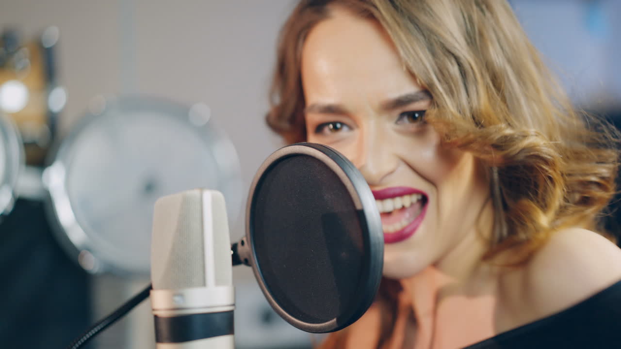 Beautiful face of a singer with bare shoulders singing into a professional modern mic in a recording studio. White and black microphone and a woman. Close-up