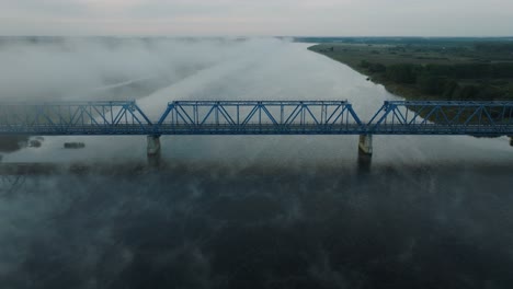 Aerial-establishing-view-of-the-steel-bridge-over-Lielupe-river-on-a-sunny-summer-morning,-fog-rising-over-the-river,-cars-driving,-wide-drone-shot-moving-forward,-tilt-down