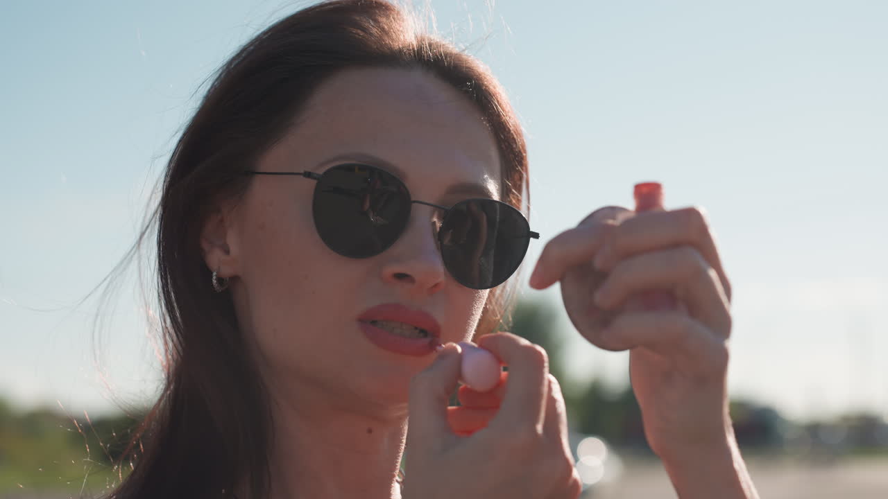 Close up of stylish lady wearing sunglasses applying lipstick while using mirror to check reflection under sunlight, highlighting beauty confidence, and personal care in urban outdoor setting