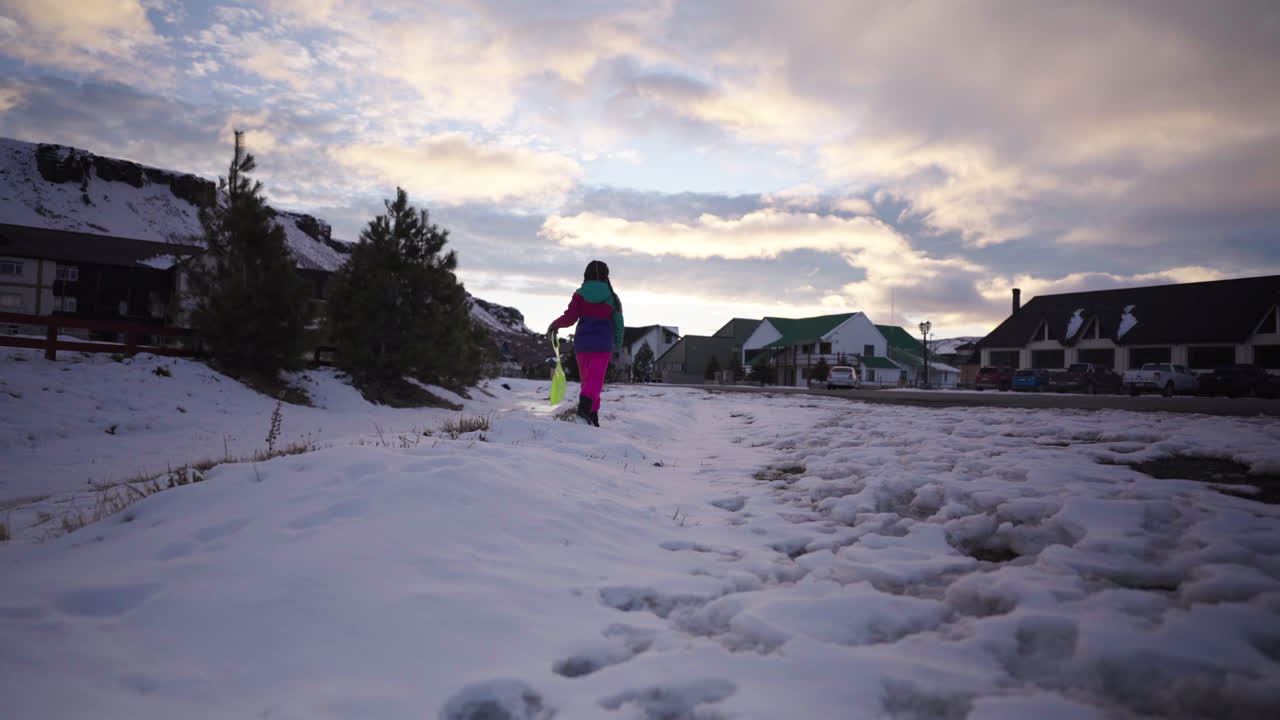 Child in colorful winter clothes walks on snow holding a green plastic sled, with village houses and mountains in the background