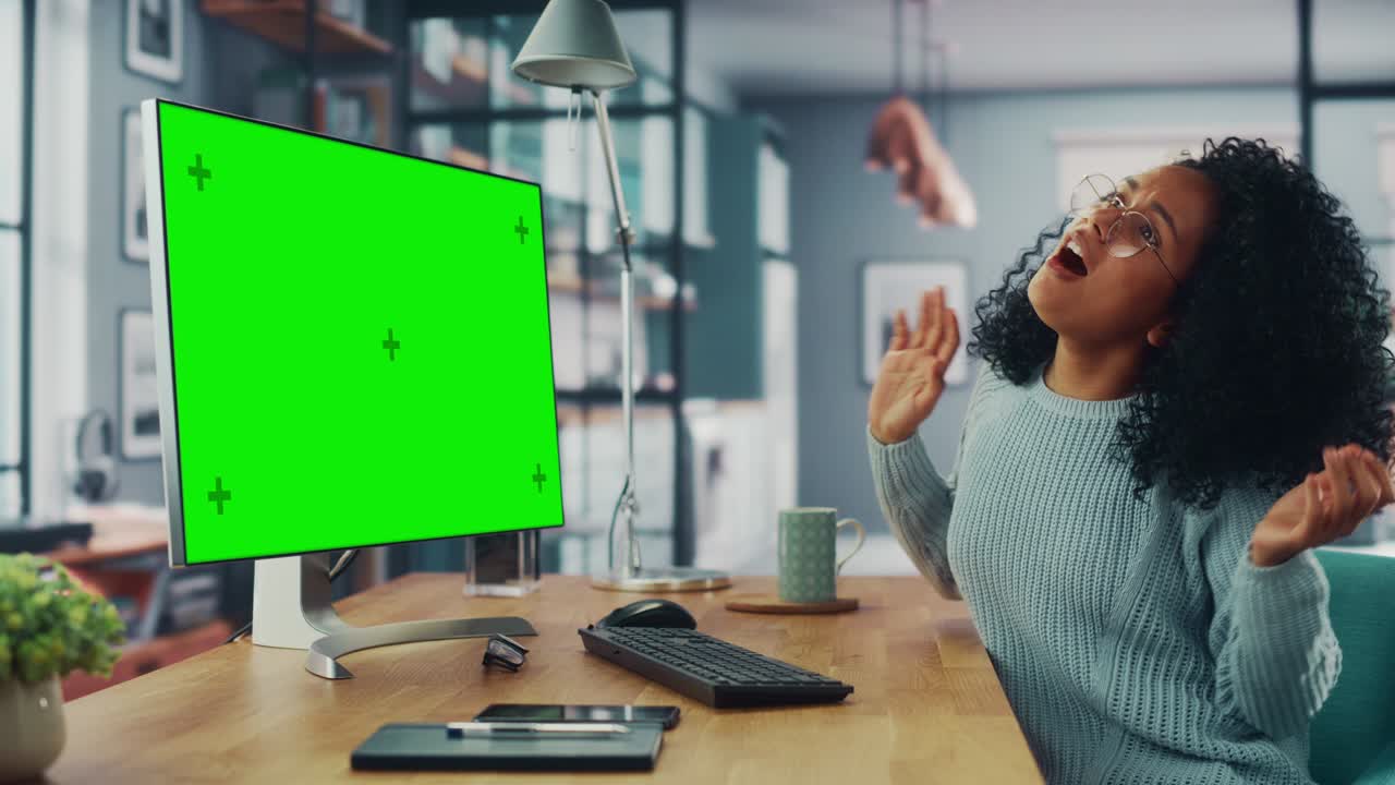 Latina Female Specialist Dancing While Sitting Next to Desktop Computer with Green Screen Mock Up Display at Home Living Room. Freelancer Female Having Fun while Remotely Working on a Project.