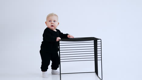 smiling baby boy stands at the grate cube. lovely blond child taps by the metal. white backdrop.