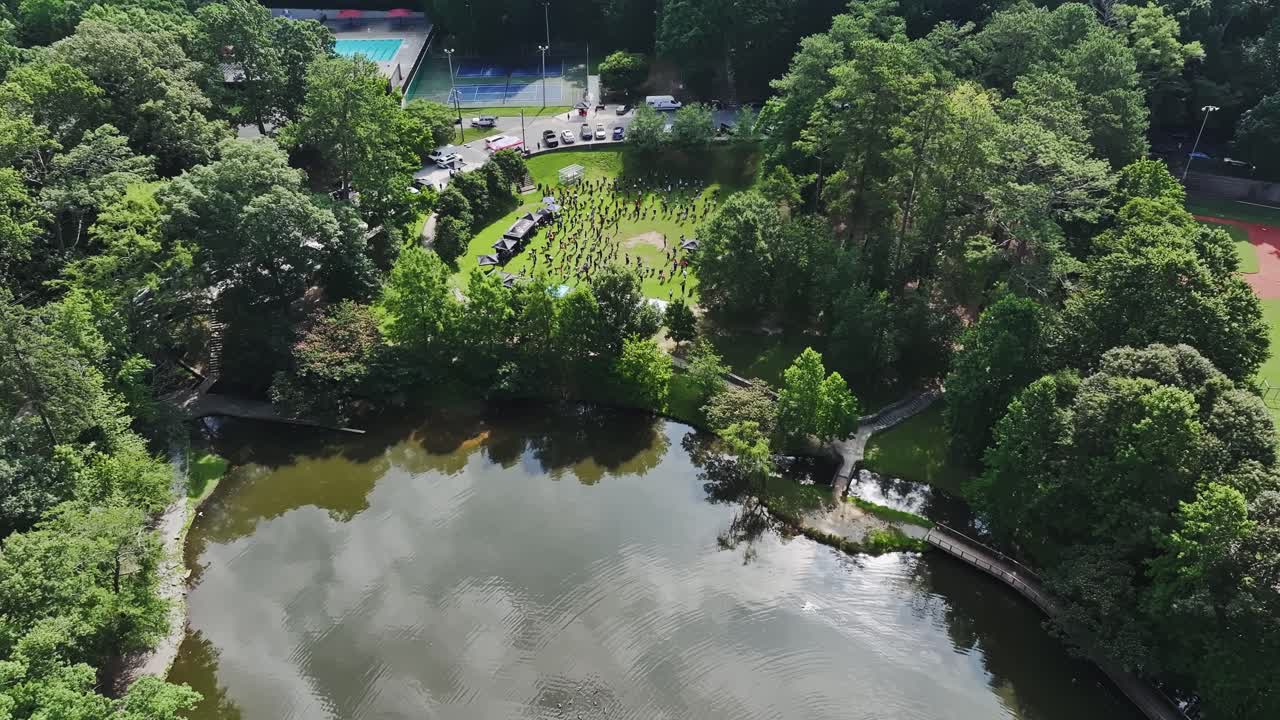 Active group of people on grass field of park near lake in summer. Suburb district of America. Town. Cars on road. Aerial landing shot