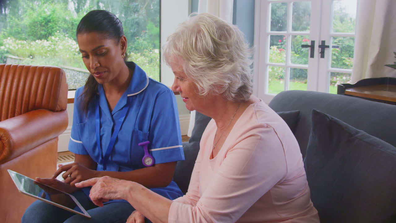 Senior Woman At Home Talking To Female Nurse Or Care Worker In Uniform Using Digital Tablet