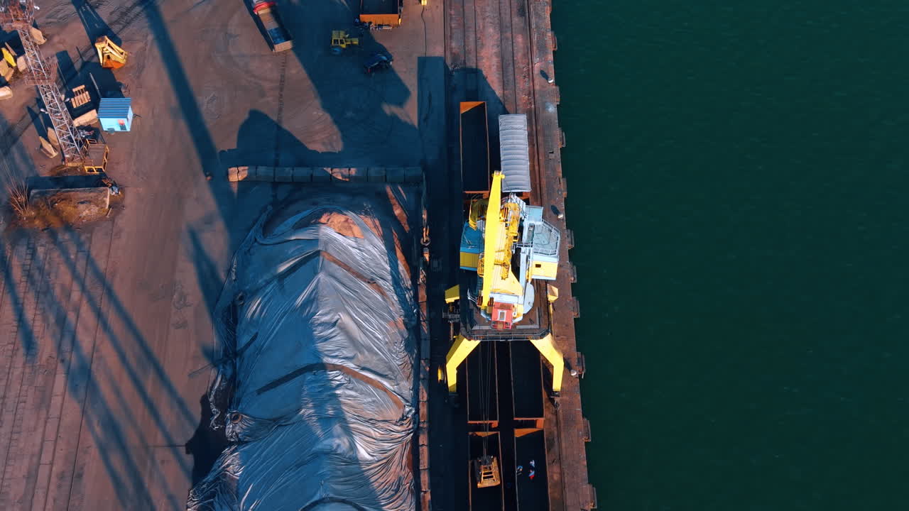 Top down aerial view of yellow harbor crane and cargo. Industrial crane moving bulk materials on a concrete pier near deep blue sea water