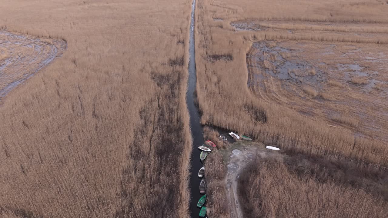 Аэрофотосъемка узкого водного канала, прорезающего обширную заболоченную местность, поросшую тростником. Несколько небольших лодок пришвартованы вдоль канала, подчеркивая присутствие человека в естественном болотном ландшафте.