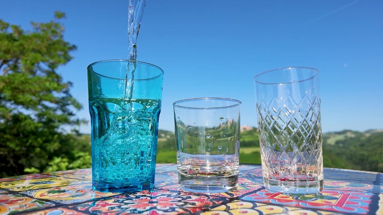 Acqua frizzante si versa in un bicchiere blu su un tavolo colorato sotto un cielo azzurro e limpido.