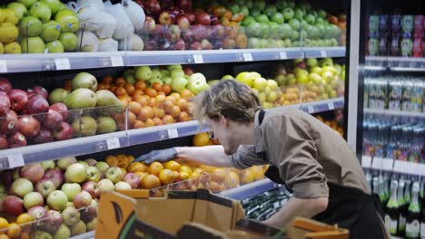 Man-worker-in-black-apron-and-gloves-stocking-the-fruits-in-supermarket.-Young-employee-at-work.-Side