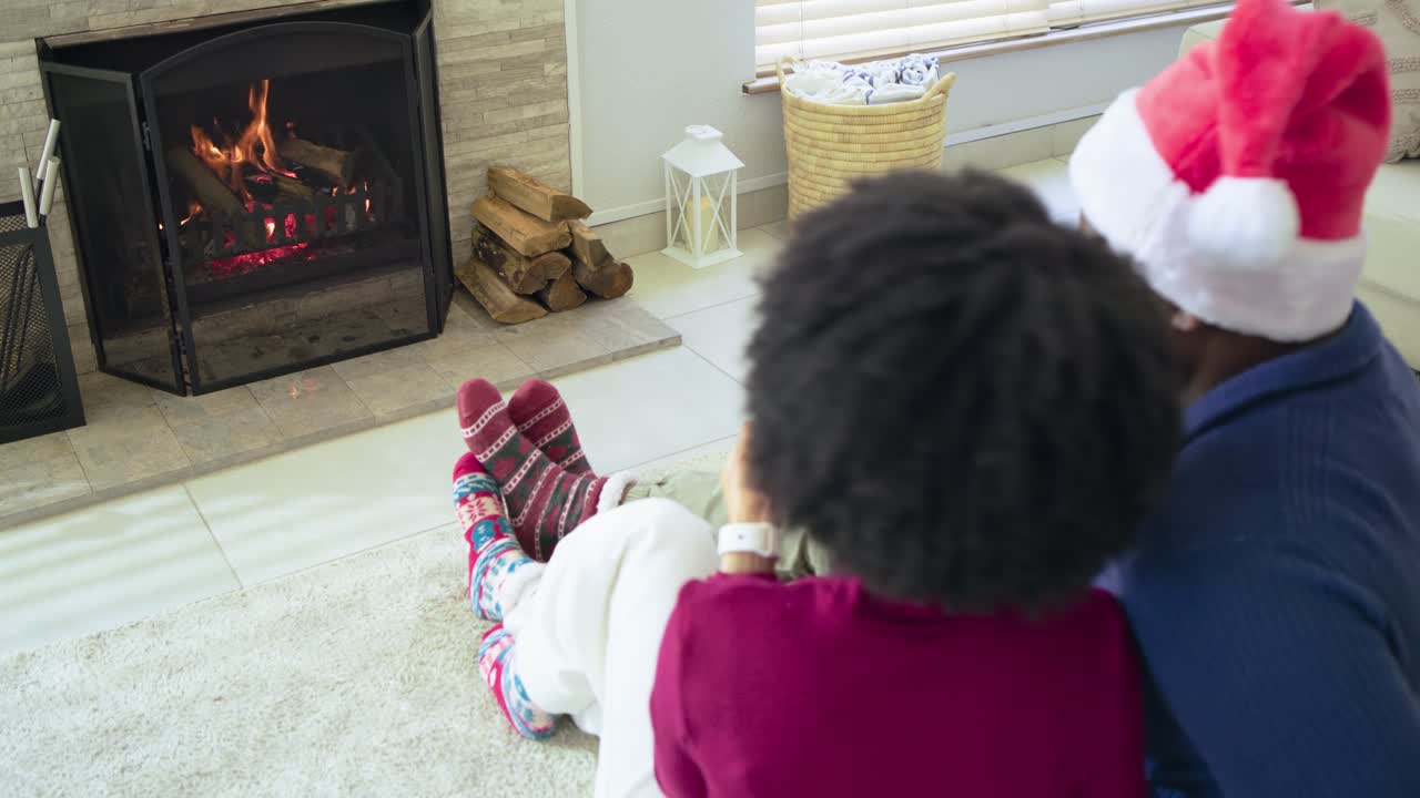 Diverse couple raising gray mugs for toast on rug before fireplace, cuddling while sipping cocoa