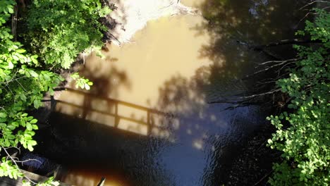 Drone-top-down-view,-wooden-pedestrian-bridge-over-shallow-stream