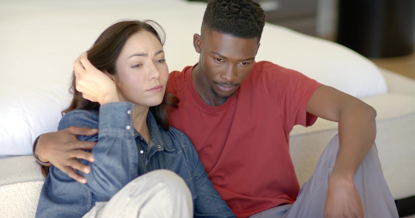 Coppia multietnica seduta accanto al letto, l'uomo che mette un braccio intorno alla donna in lacrime, che si appoggia a lui per conforto.