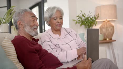 Happy-senior-biracial-couple-sitting-on-sofa-and-using-laptop,-unaltered,-in-slow-motion