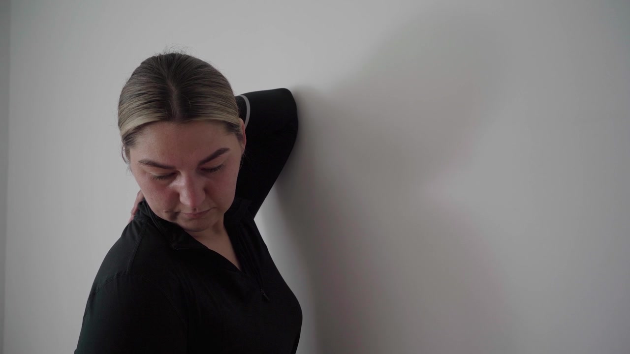 Mujer con ropa deportiva oscura realiza un estiramiento de tríceps y hombros por encima de la cabeza contra una pared en blanco para aliviar la tensión de la parte superior del cuerpo.
