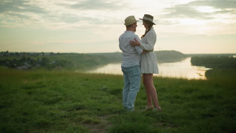 two happy people hold each other while dancing on a grassy hill beside a tranquil lake. the man is dressed in a white shirt, hat, and jeans, while the woman wears a white dress and black hat