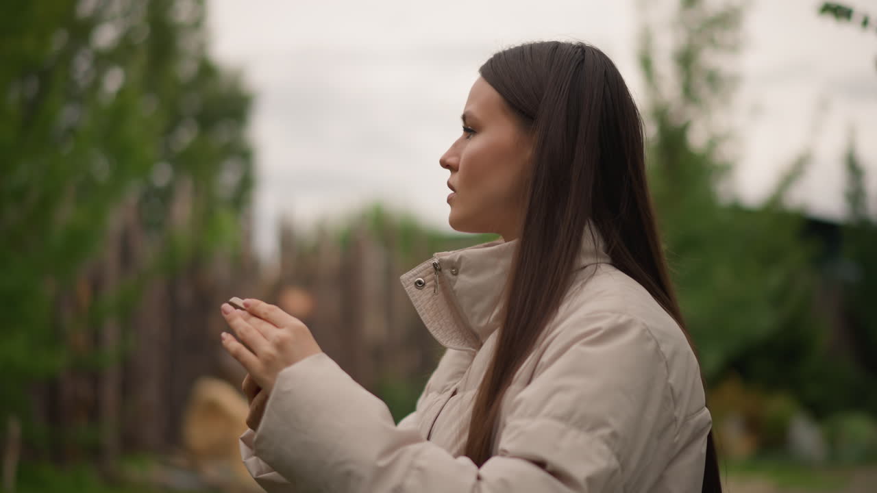 Mujer caucásica de perfil fotografiando con un smartphone, chaqueta acolchada beige y pelo largo trenzado, valla de madera y bokeh de patio trasero, luz suave y nublada, expresión concentrada, encuadre de toma vertical