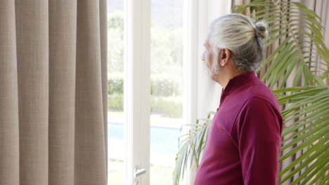 thoughtful biracial senior man with white beard looking out of sunny window, copy space, slow motion