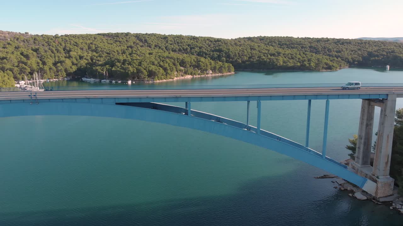 Aerial view lonely car driving fast on a concrete bridge over the ocean in a holiday paradise with wild nature and yacht parking on the beach
