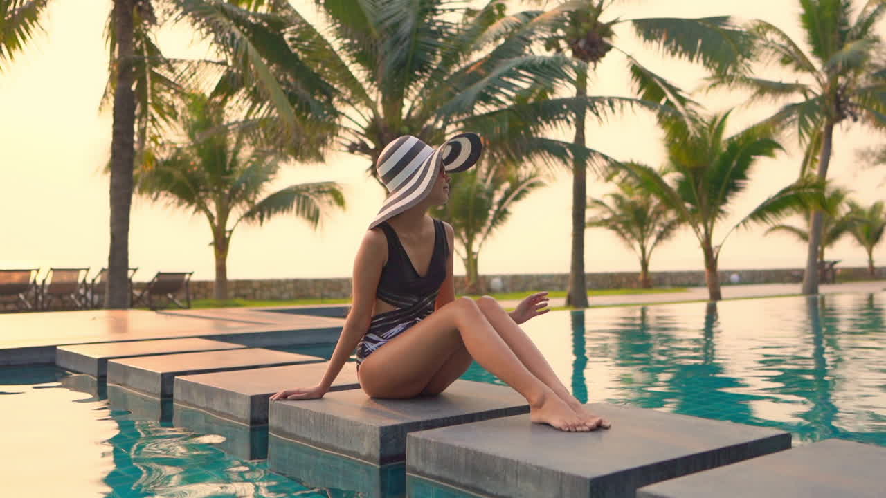 Sexy Asian lady sits near a swimming pool on floating cubes wearing a large black and white hat with sunglasses and swimwear in Exotic resort in Thailand on sunset
