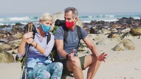 senior hiker couple wearing face masks with backpacks and hiking poles talking sitting on rock