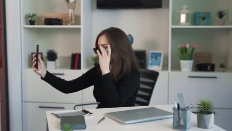 The-young-woman-is-sitting-at-a-desk-in-an-office,-looking-into-the-camera-of-her-phone-and-trying-on-sunglasses