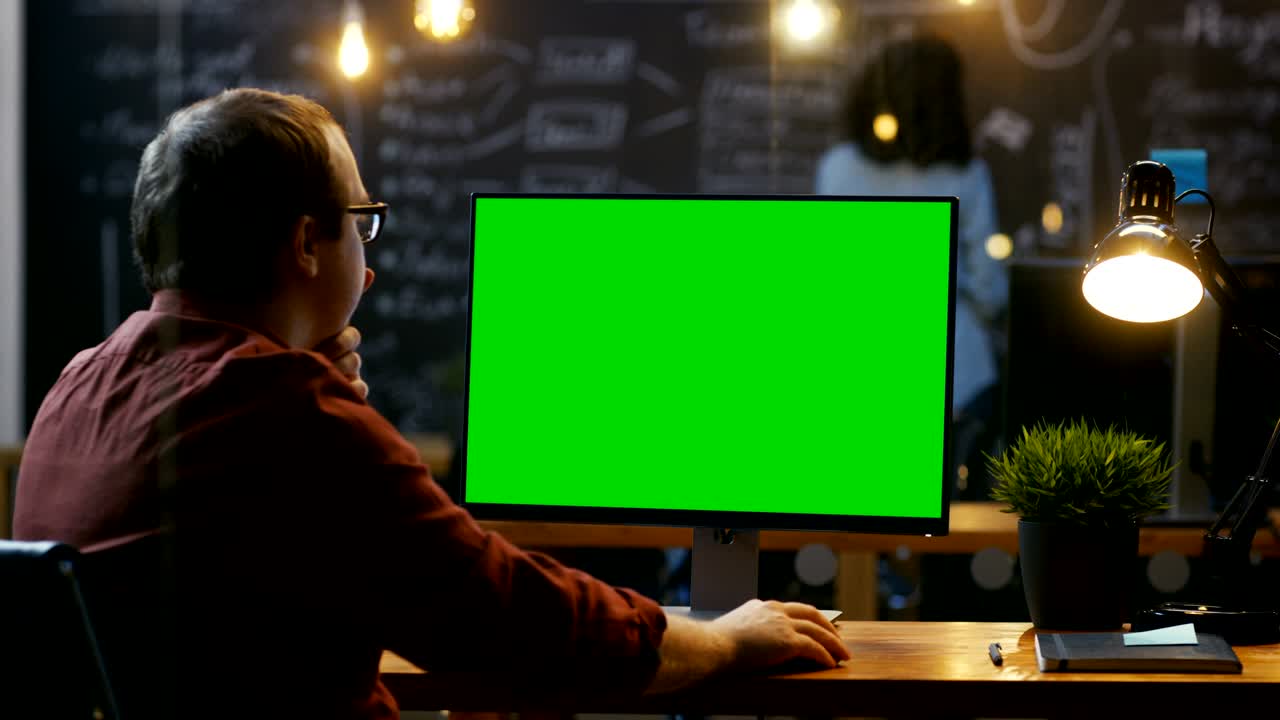 Office Employee at His Desktop Works on a Mock-up Green Screen Personal Computer. Over the Shoulder Footage. His Colleague works in the Background. Creative Office Evening.