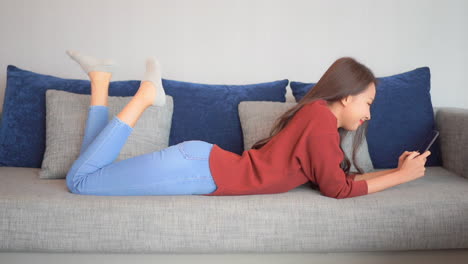 young woman laying on stomach using smartphone in living room on sofa checking online profile or dating app