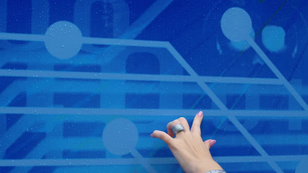 한 여자의 손은 방울로 된 캔버스를 가로질러 흐르고, 파란 바탕에 손가락으로 선을 그려서 기묘한 미래의 패턴을 형성합니다. 손가락에는 은 반지가 있습니다.