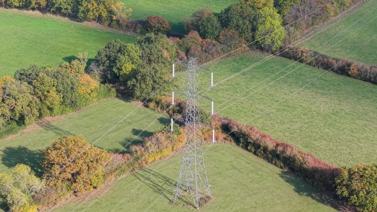 Aerial drone orbit around electricity pylon in Hertfordshire, UK. October sunlight casts long autumn shadows over the trees and grass, highlighting the rural landscape and power infrastructure below