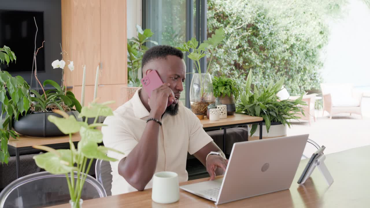 African man receiving call on pink smartphone, using tablet and laptop for work at wooden table