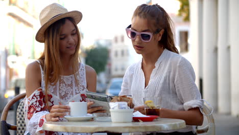 Two-female-friends-read-a-guidebook-outside-a-cafe,-Ibiza