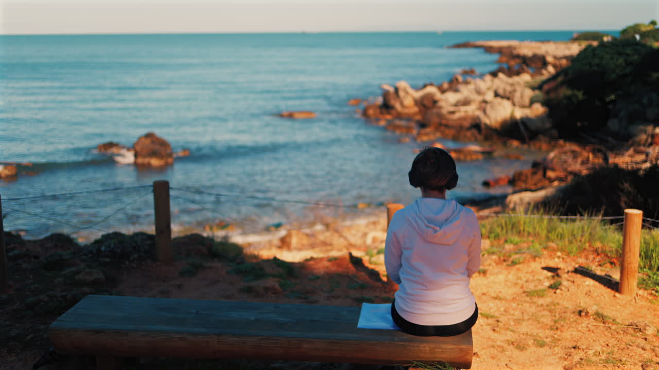 Woman with headphones on her head sitting on a wooden bench with a view of the sea