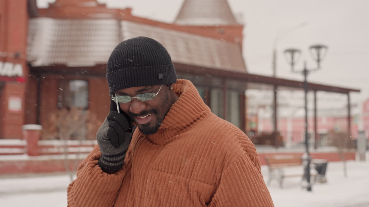 Uomo di colore su una strada innevata che parla al telefono, indossa un cappotto teddy marrone, berretto nero, guanti e occhiali, che cammina davanti a vetrine di mattoni rossi sotto una leggera nevicata, espressione pensierosa mentre controlla lo schermo.