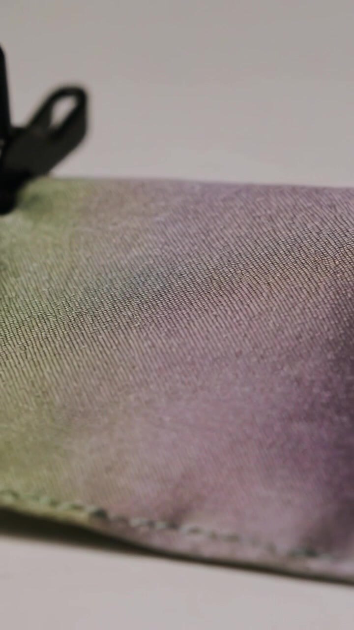 Video vertikal: Menampilkan kantong dengan anyaman diagonal yang berubah warna di atas meja saat cahaya bergerak, dengan detail tarikan ritsleting.