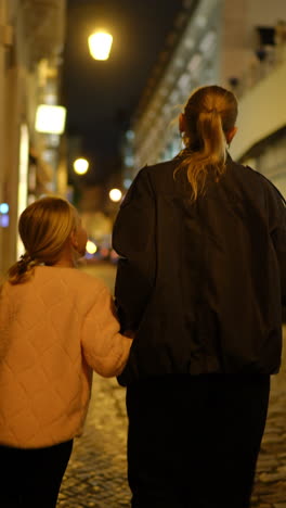 loving mother guiding young daughter while walking hand in hand along narrow cobblestone street, streetlamps casting soft shadows during evening stroll through historic european urban neighborhood