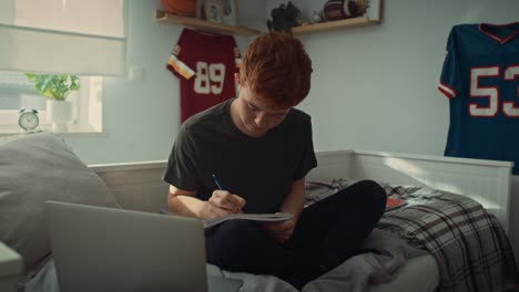 caucasian teenage boy sitting on bed and making notes from laptop.