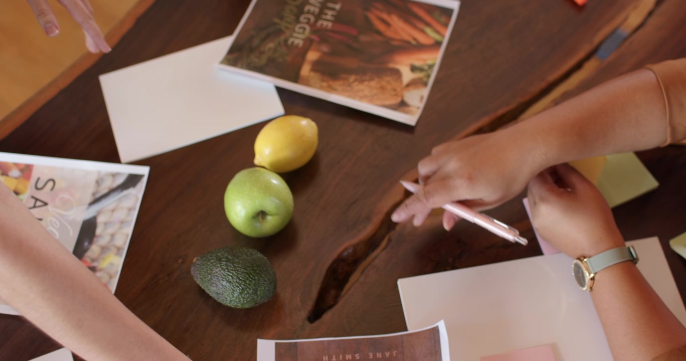 Due donne che pianificano uno shooting di ricette su un tavolo con bordo naturale, disponendo un libro di ricette, frutta, appunti e spazio per il testo.