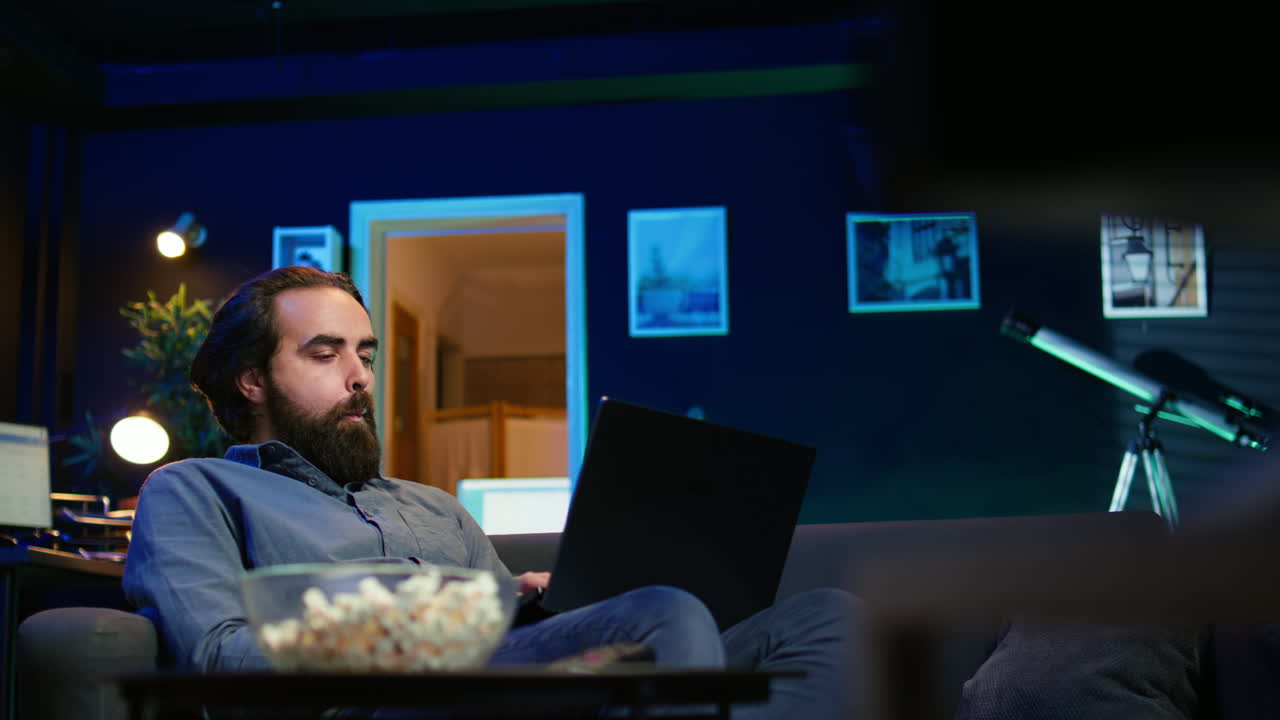 hombre trabajando en una computadora portátil en casa con un programa de televisión jugando en el fondo