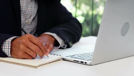 mid section of businessman writing on notepad while working on laptop