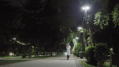 peaceful night scene in beautiful park with lady walking in white coat along illuminated path, street lamps glowing above, and silhouettes of people visible in distant background