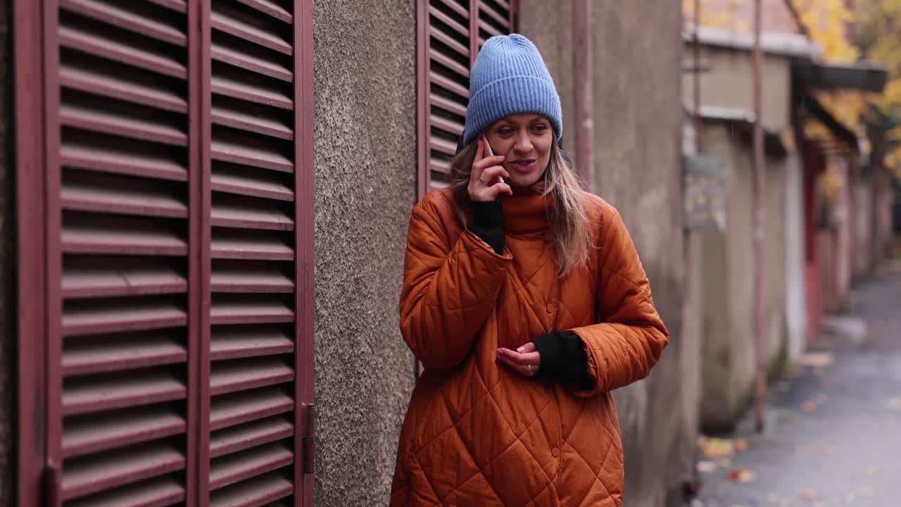Woman in orange jacket and blue hat happily talks on phone
