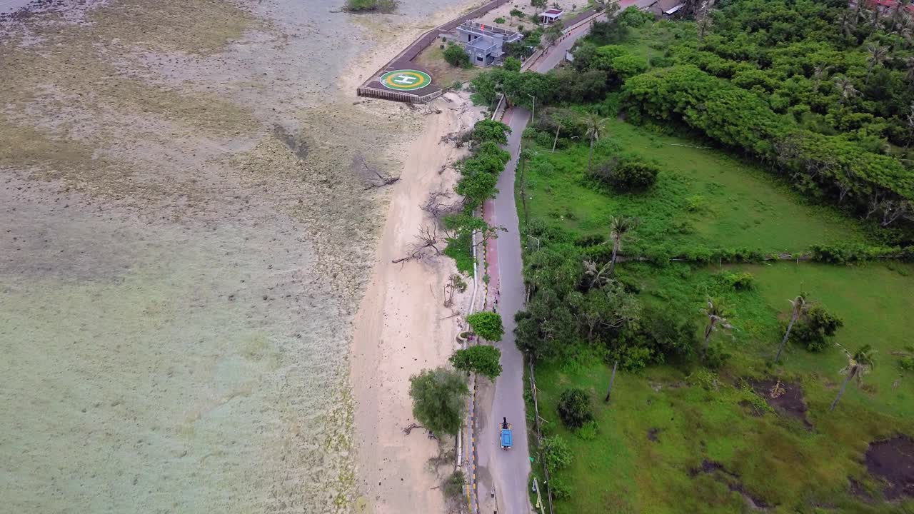Zoom in shot over a coastal road and lush green fields beside the shoreline at Gili Trawangan on a clear day